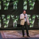 comedian Shaun Eli on stage at Mamaroneck's Emelin Theatre in front of Ivy logo banner, standing on an Oriental rug
