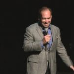 Clean stand-up comedian Shaun Eli on stage at a New York theatre, smiling down at the audience